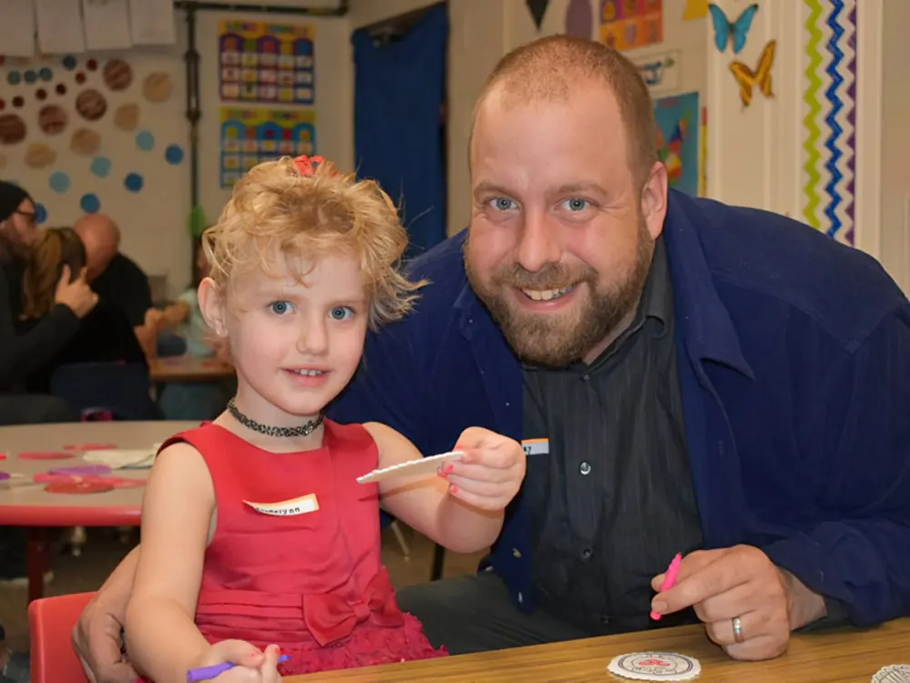 A smiling man and a little girl in a red dress at a classroom craft table