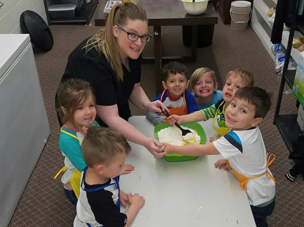 A teacher and five children in aprons mixing something in a big bowl at a classroom table