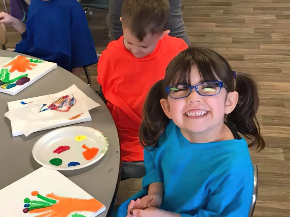 A smiling girl in glasses at a table covered in finger paintings