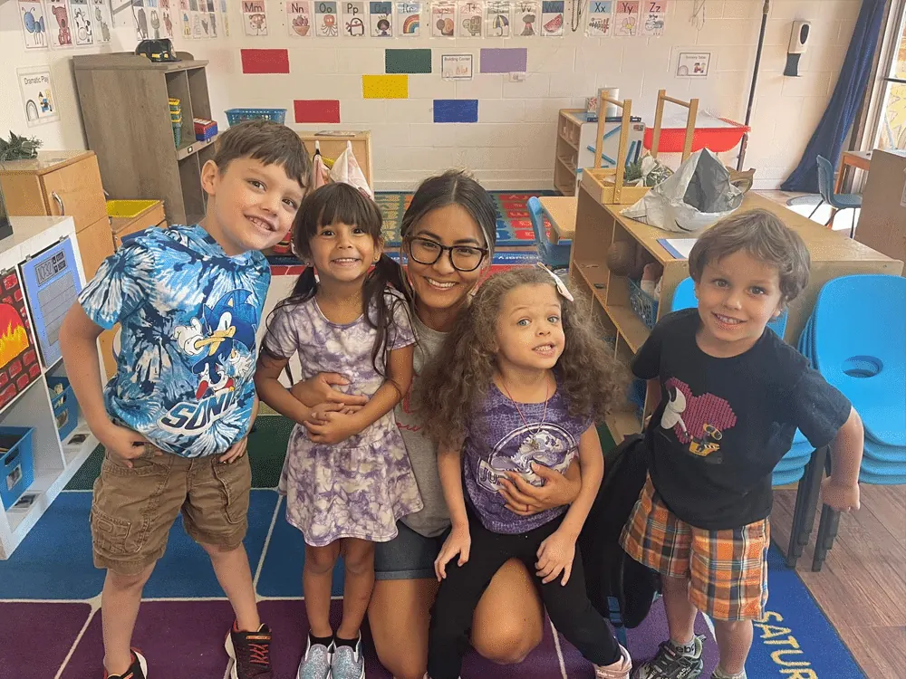 A teacher kneeling with five smiling children in a classroom