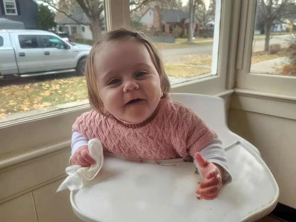 A smiling baby girl in a pink sweater in a high chair by a window