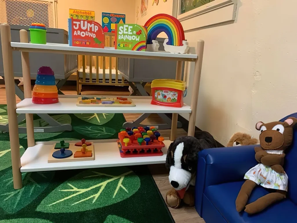 A classroom corner with toy shelves, picture books, a rainbow and stuffed animals on a leaf-patterned rug
