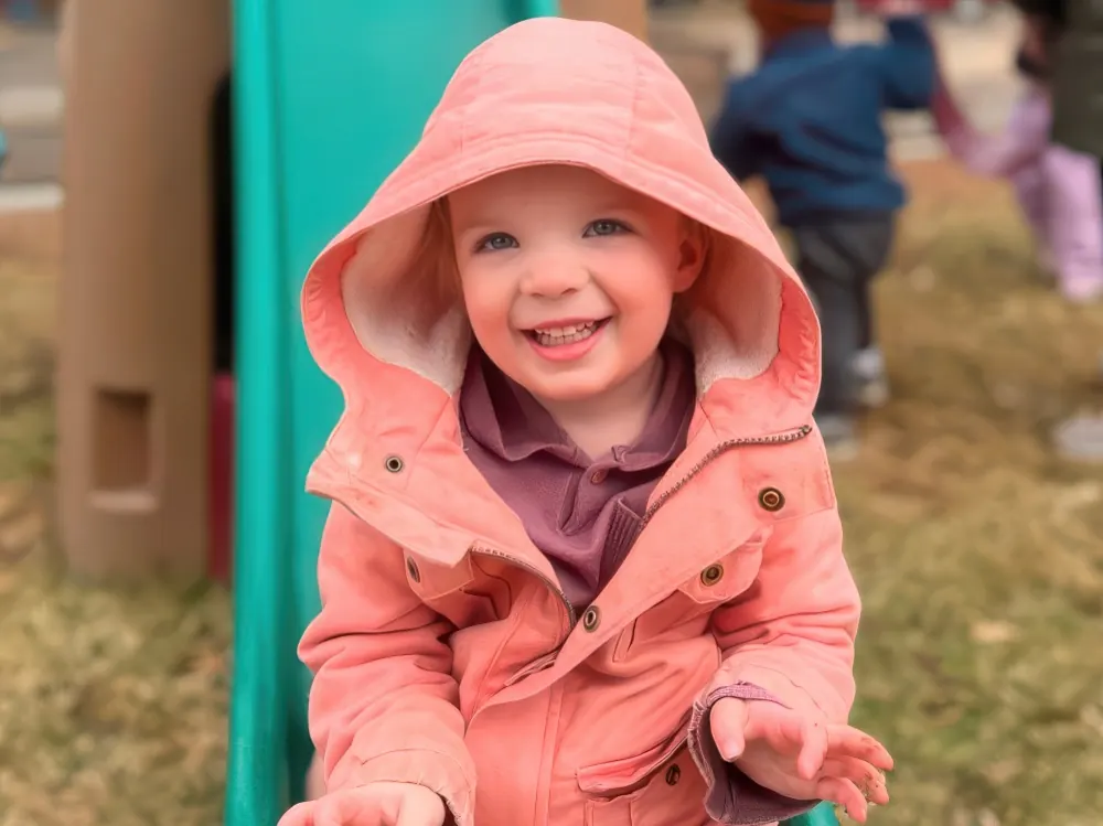 A smiling toddler in a coral hooded jacket on a playground