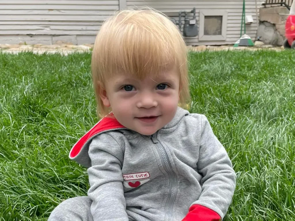 A smiling blond baby in a gray zip-up sitting on the grass