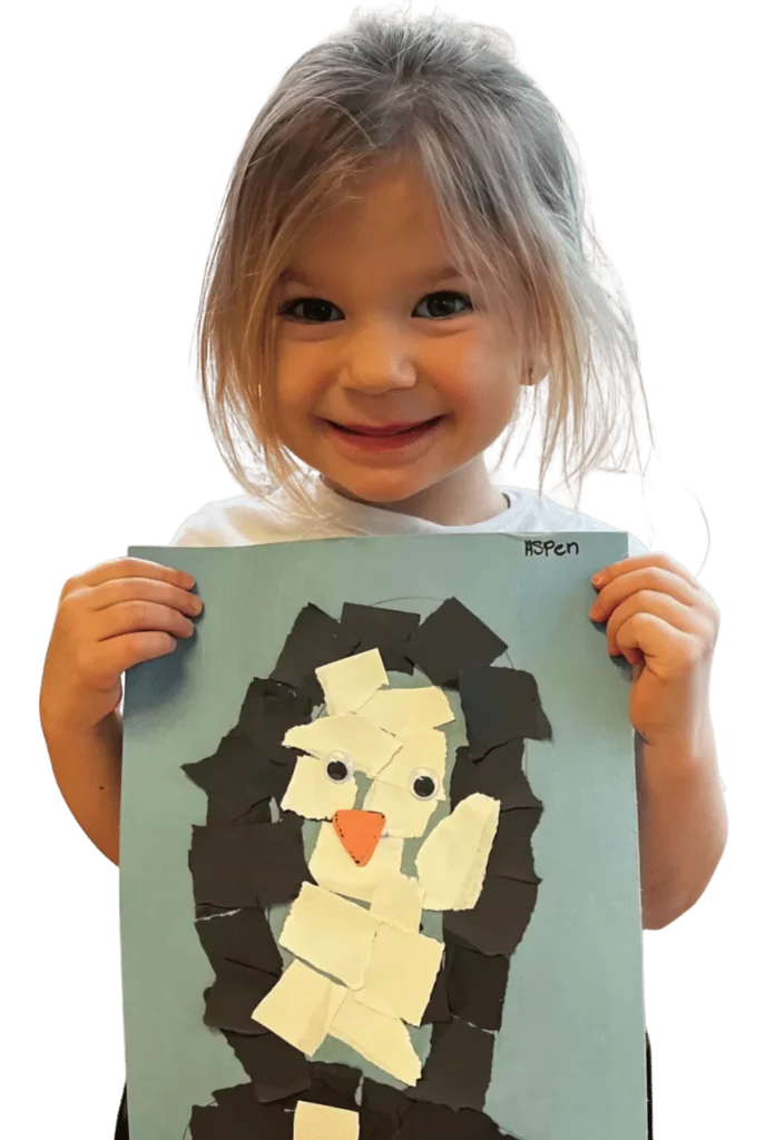 A smiling girl holding up her torn-paper penguin collage