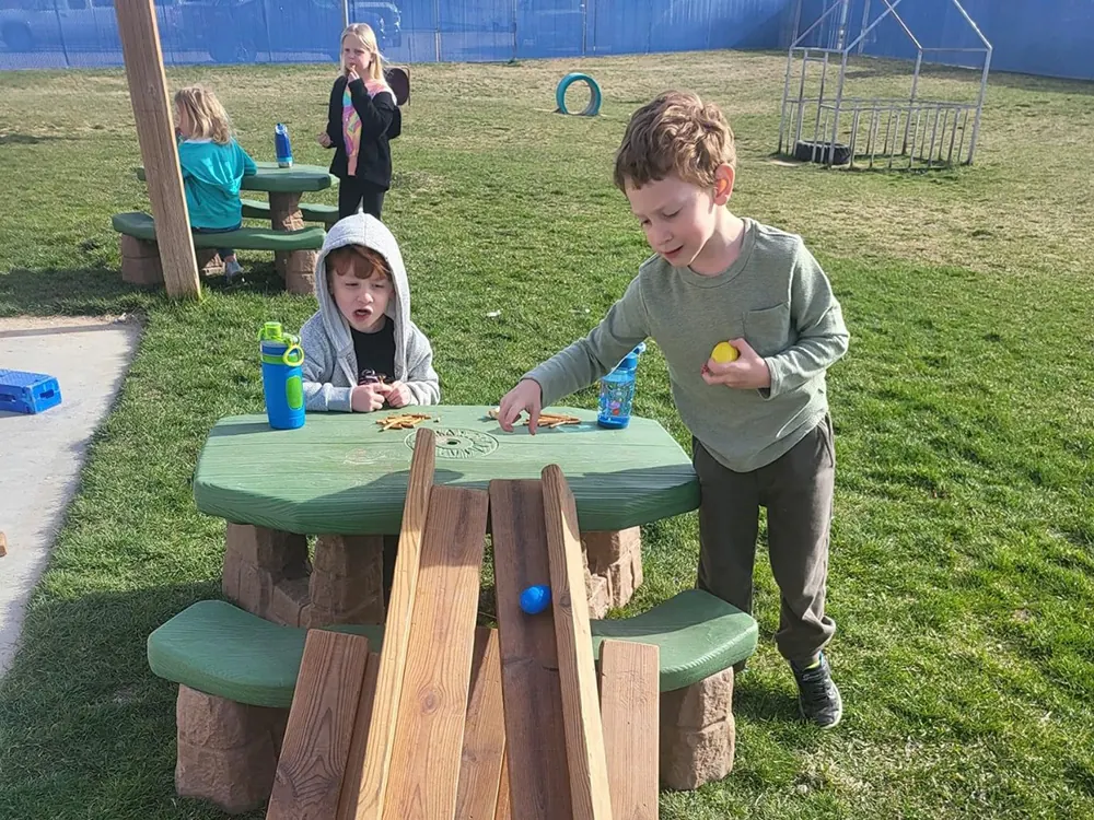 Children at a green picnic table on the playground, rolling balls down a wooden ramp