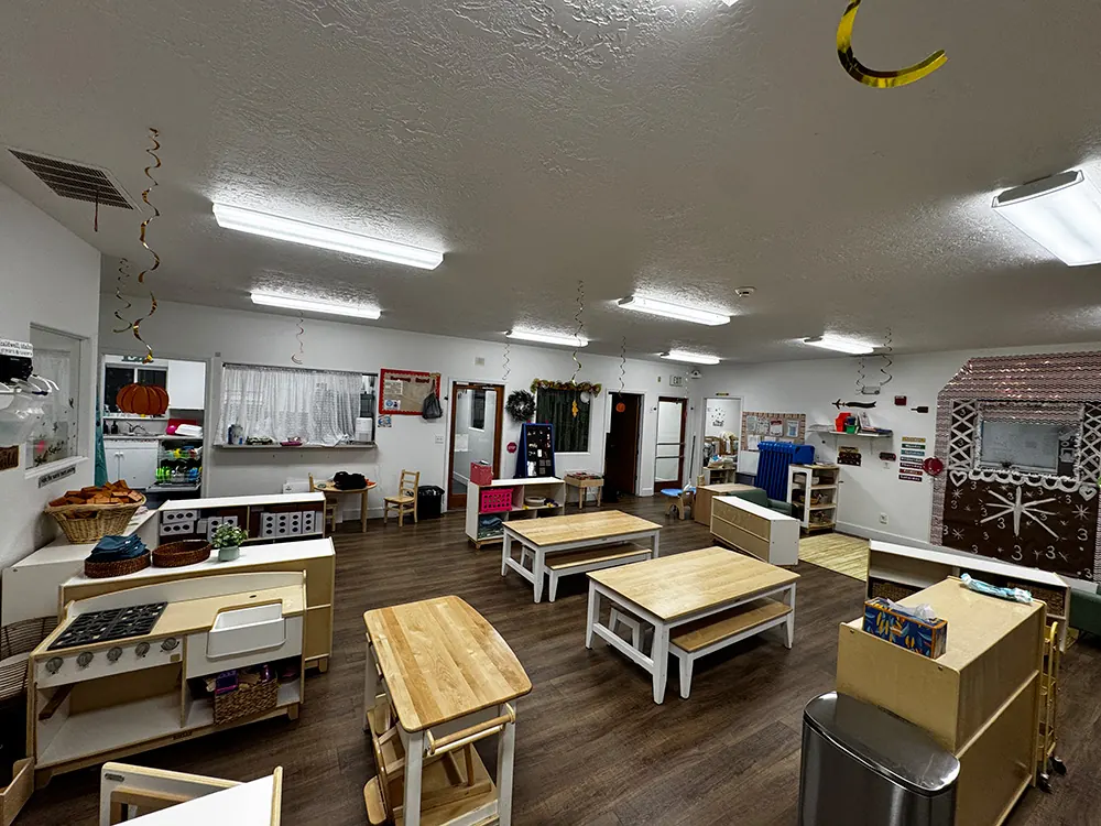 A large, bright classroom with child-size tables, a play kitchen and low shelves