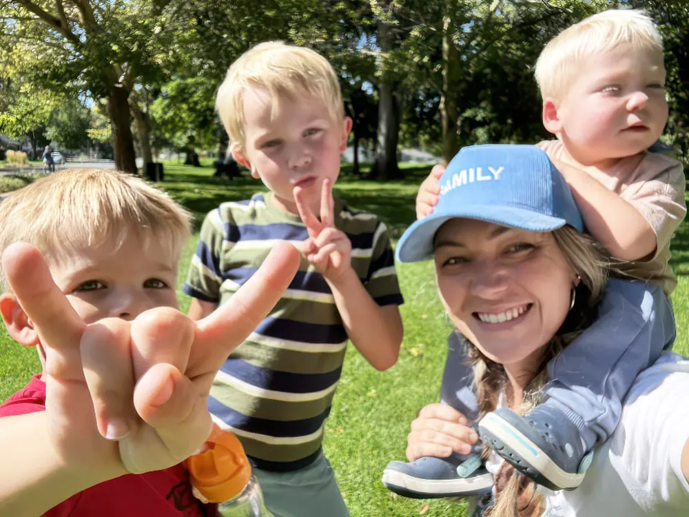 A teacher carries a toddler on her shoulders beside two boys making peace signs in a park
