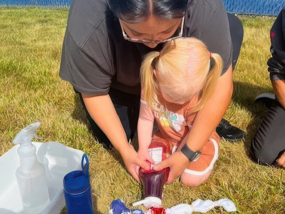 A teacher helps a little girl squeeze dye onto a shirt on the grass