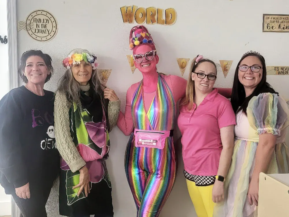 Five teachers in bright, playful costumes smiling together in a classroom