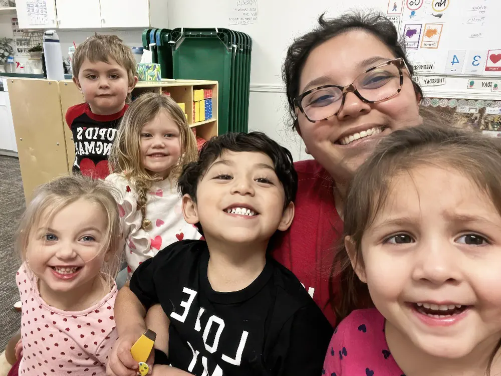 A smiling teacher takes a selfie with five happy children in a classroom