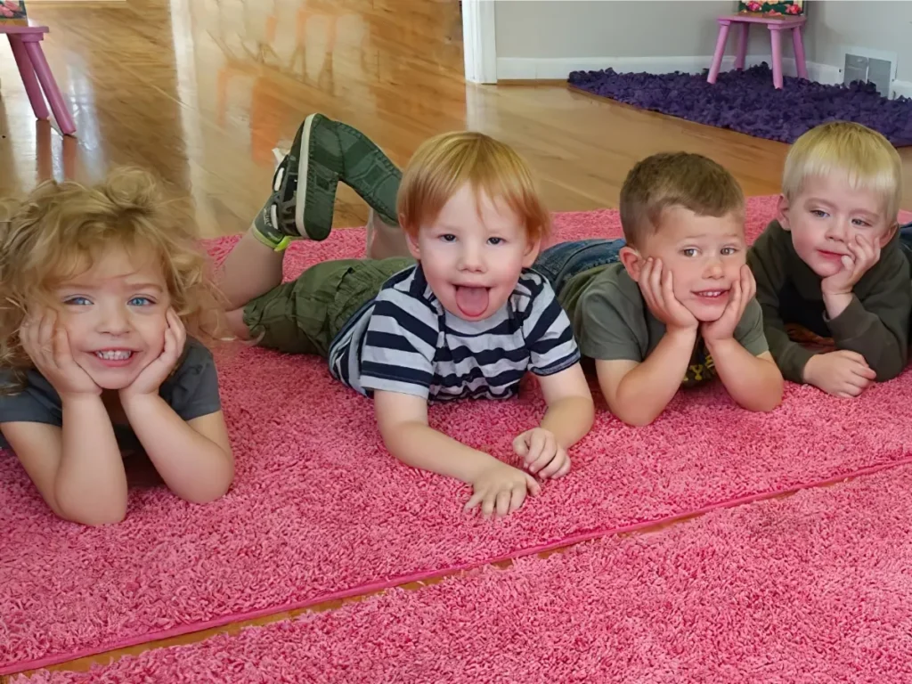 Four toddlers lying on a pink rug with their chins in their hands, one sticking out his tongue
