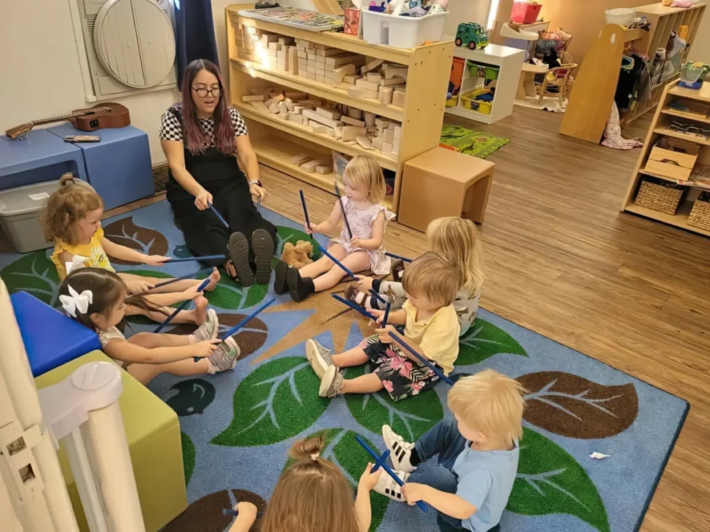 A teacher leading seated toddlers in a rhythm-stick activity on a leaf-patterned rug