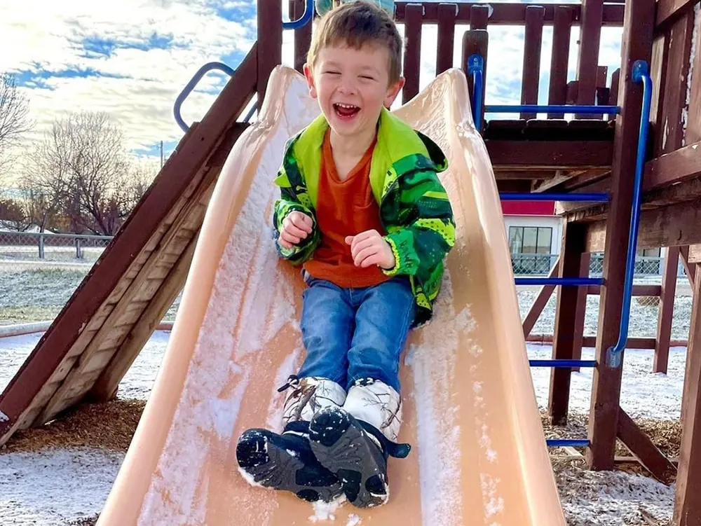 A laughing boy in a green jacket and snow boots coming down a slide on a wooden play structure