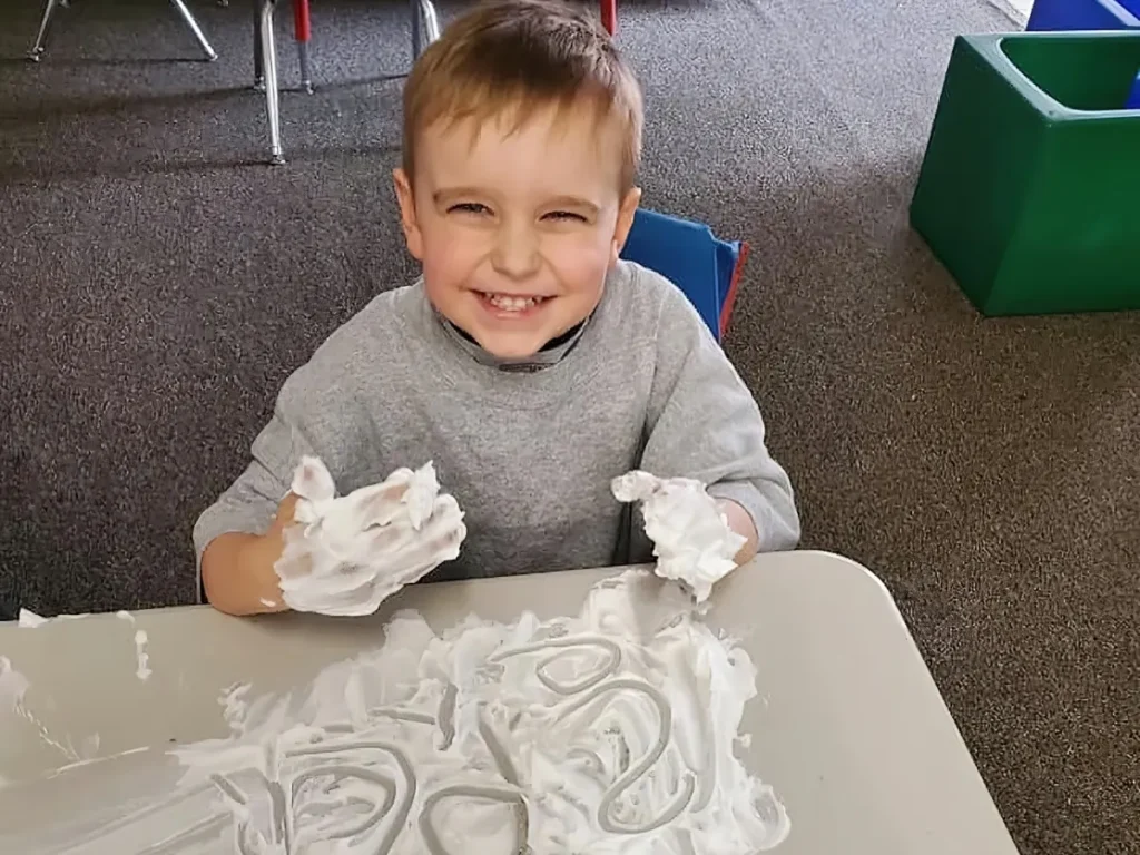 A grinning boy with shaving-cream-covered hands at a table smeared with foam