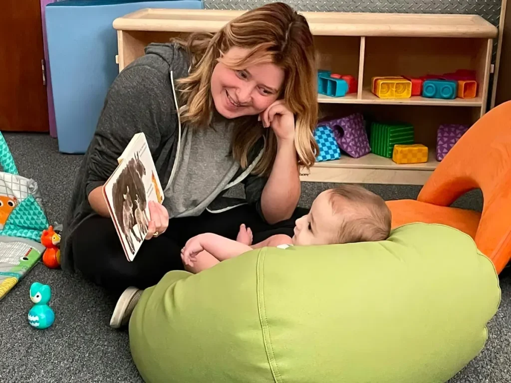 A teacher sitting on the floor reading a board book to an infant on a green beanbag