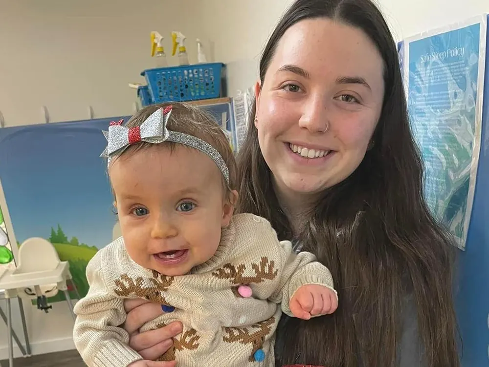 A smiling teacher holding a baby girl in a reindeer sweater and a sparkly bow headband