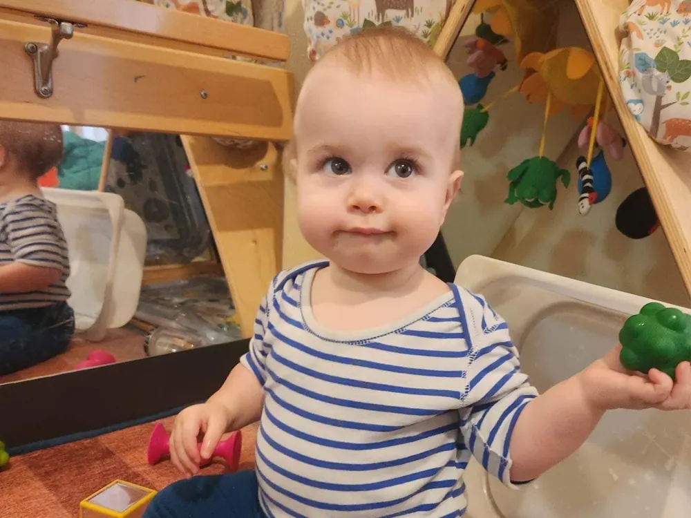 A baby in a striped shirt standing at a sensory table, holding a green ball