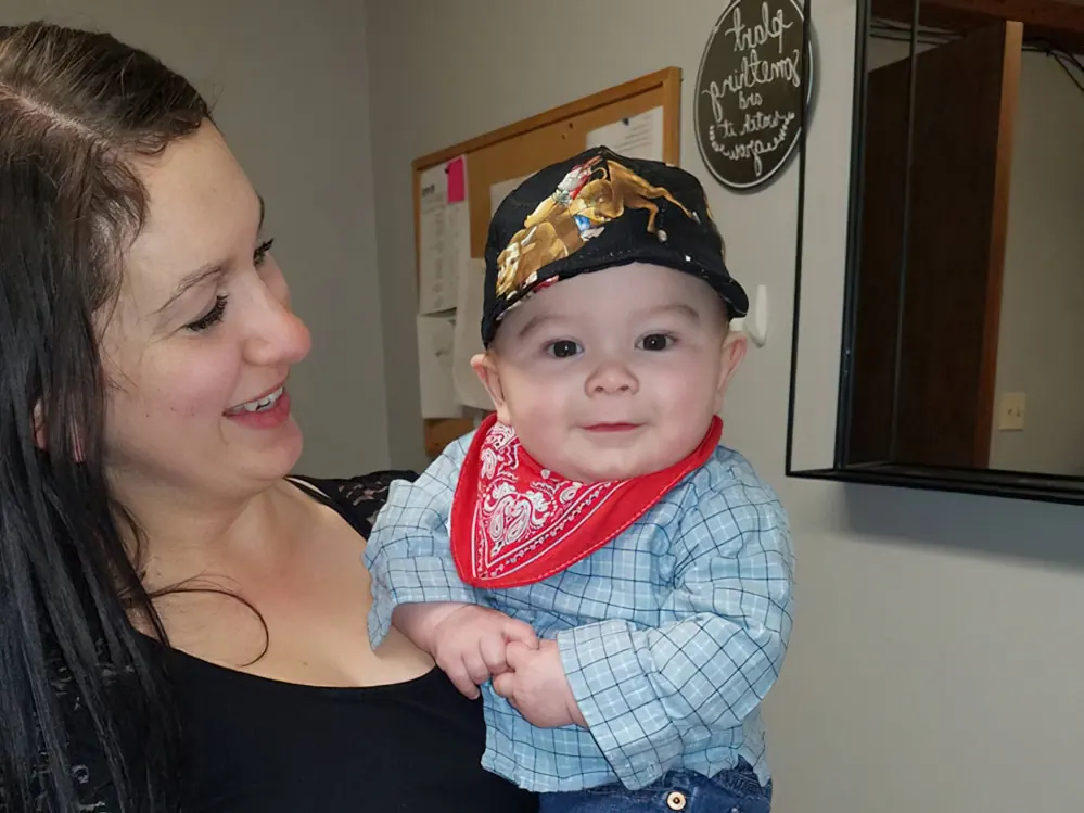 A smiling mother holding her baby boy, who wears a cap, a red bandana and a checked shirt