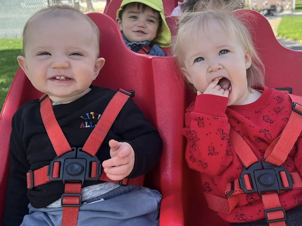 Three babies buckled into a red multi-seat stroller outdoors