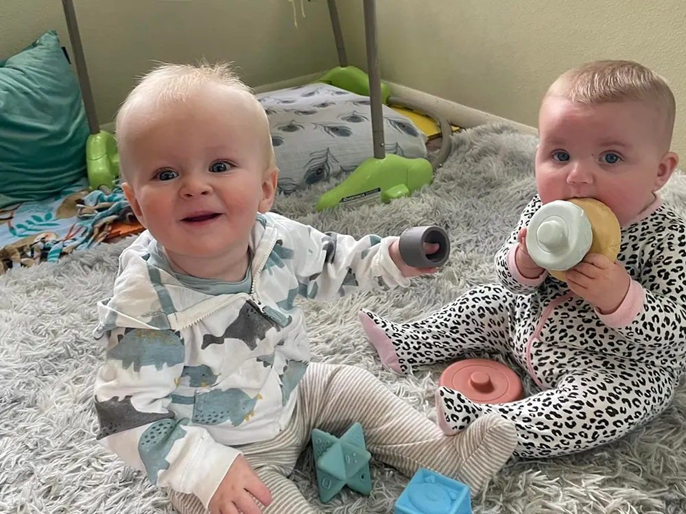 Two babies in footed pajamas sitting on a fluffy rug, one holding a bottle