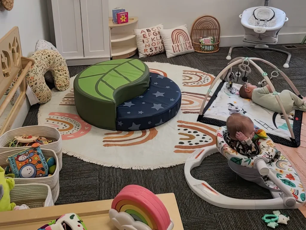 Babies on a play mat and in a bouncer in a bright infant room with a leaf-shaped floor cushion and a rainbow rug