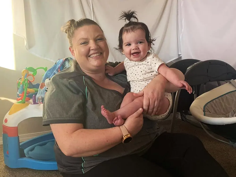A smiling teacher holding a laughing baby girl with a topknot on her lap