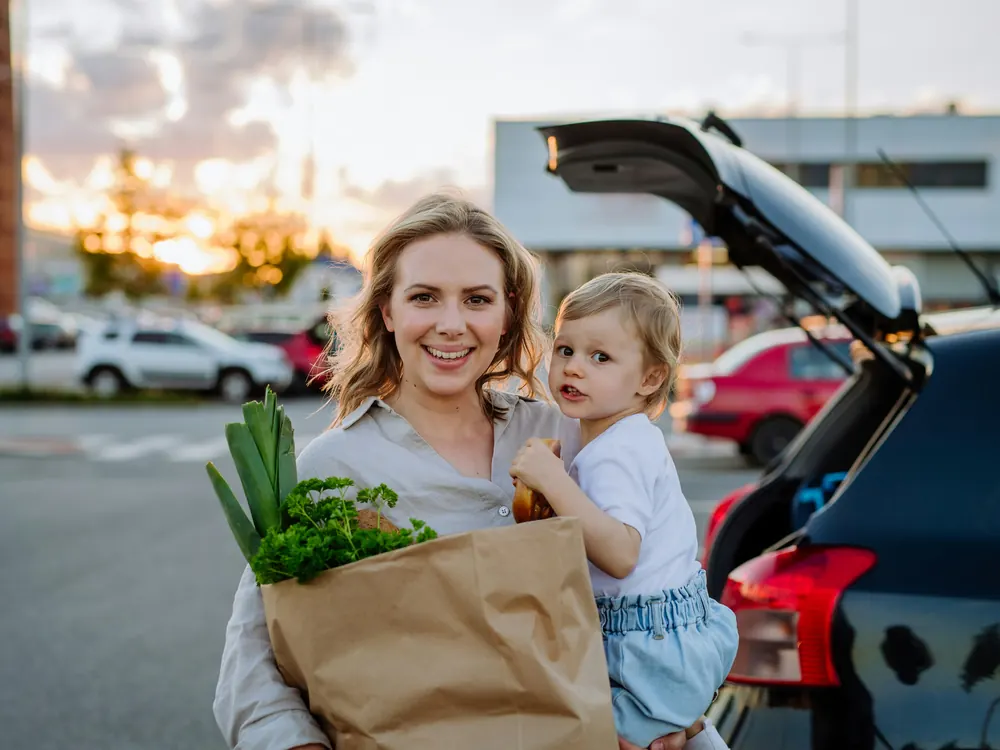 A smiling mother holding her toddler and a bag of groceries beside her car