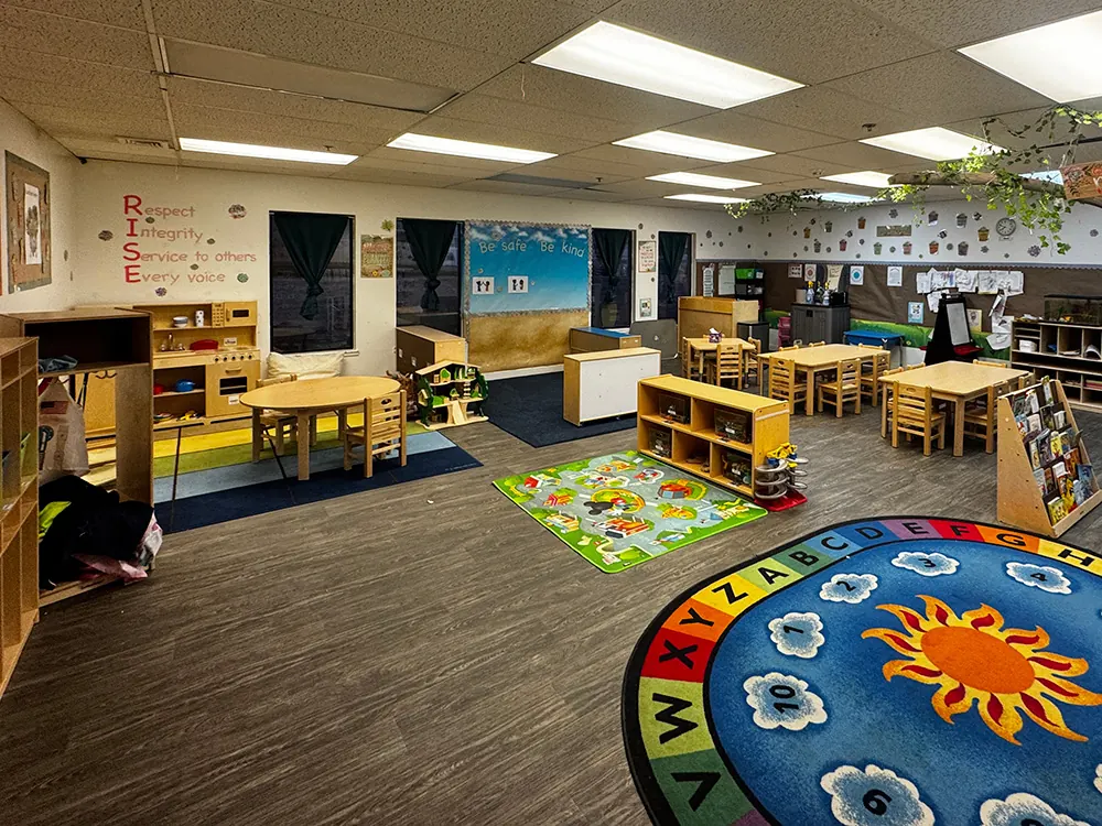 A large, bright classroom with a round alphabet-and-sun rug, low shelves and child-size tables