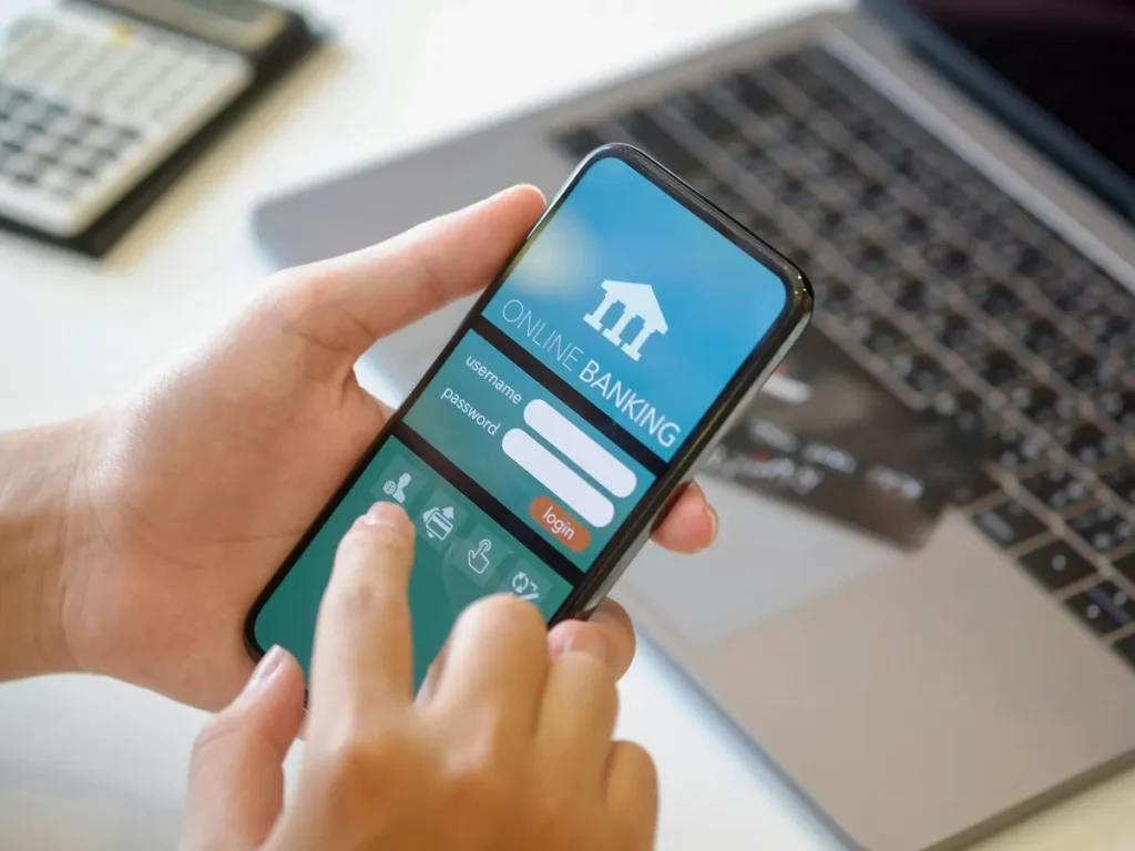 Hands holding a phone with an online banking app open, a laptop behind it