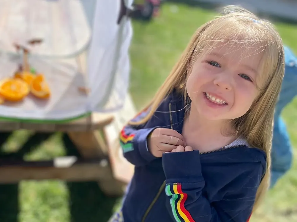 A smiling girl with long blond hair outdoors, her hands clasped at her chest