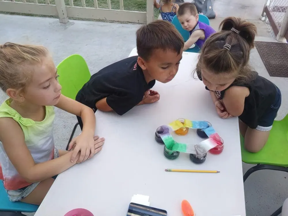 Three children leaning over a table to watch a color-mixing experiment on paper towels