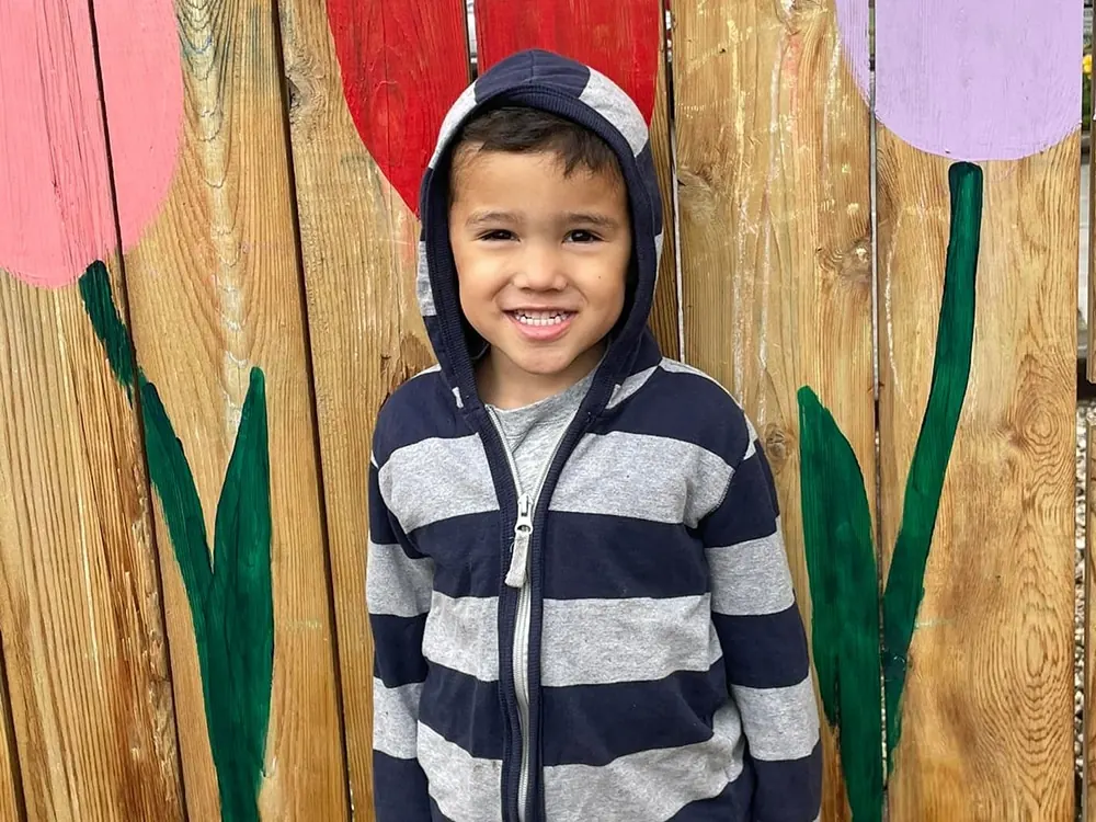 A smiling boy in a striped hoodie in front of a painted wooden fence