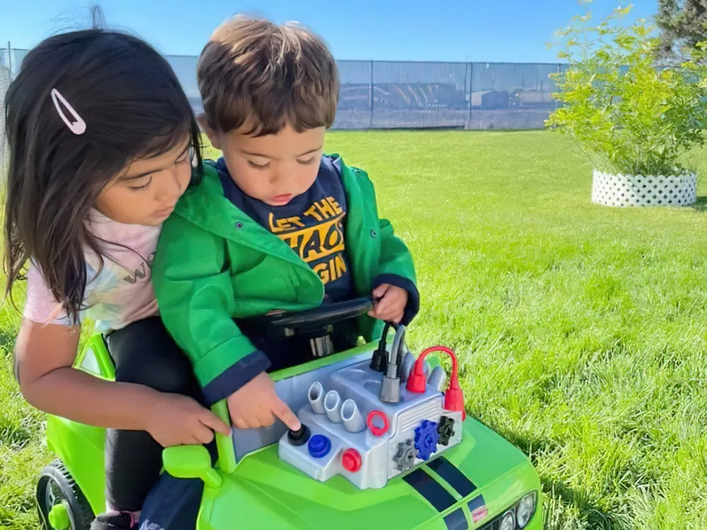 A girl helping a toddler boy steer a toy car on the grass