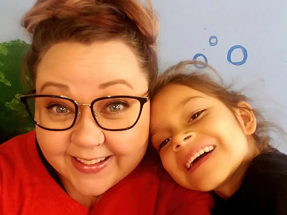 A smiling teacher in glasses cheek to cheek with a laughing girl