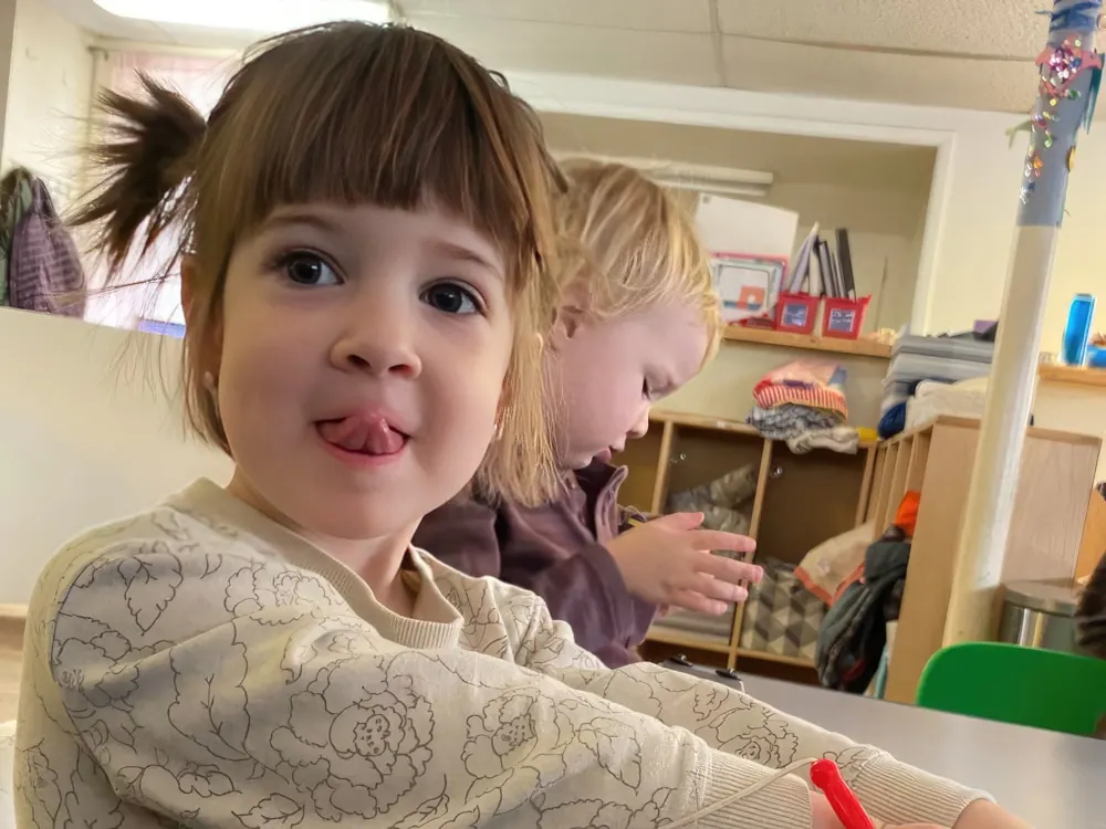 A girl with bangs sticking out her tongue at a classroom table, another child behind her