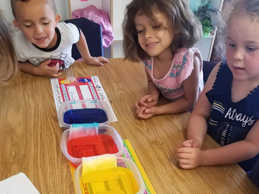 Three children watching a color-mixing experiment, with red, blue and yellow water creeping along paper towels between tubs