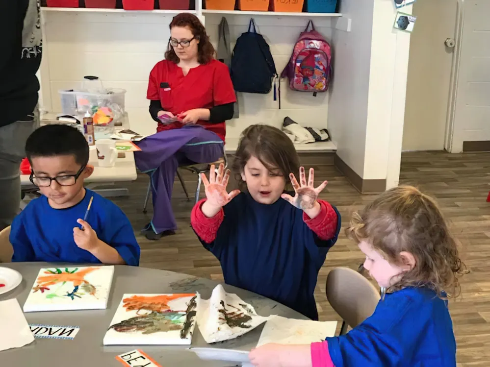 Children in blue smocks painting at a table while a teacher in red scrubs sits behind them