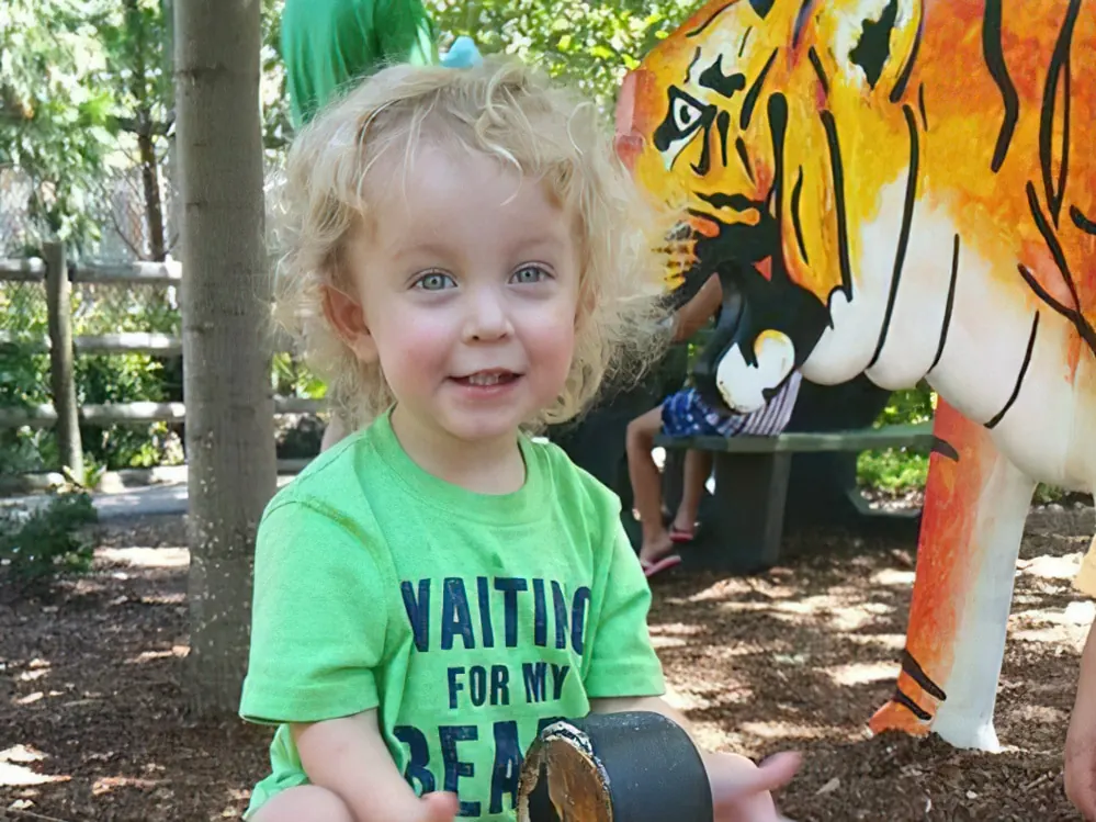 A curly-haired toddler in a green T-shirt at the zoo beside a painted tiger sculpture
