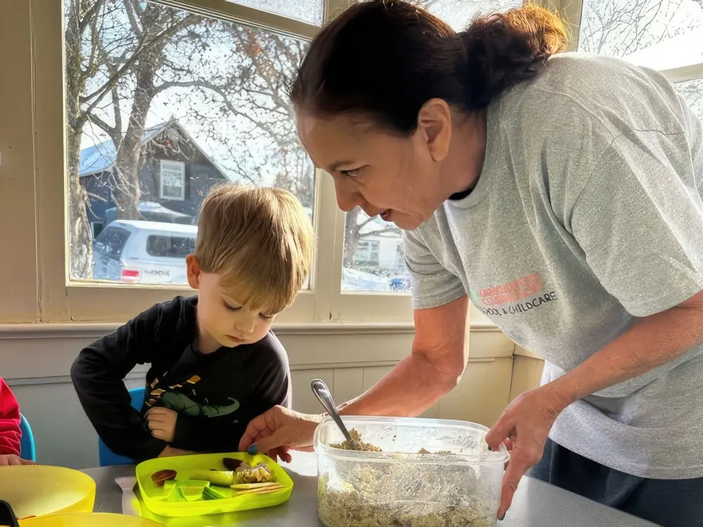 A staff member in a Kangaroo Clubhouse T-shirt serving food to a boy at a table by a window