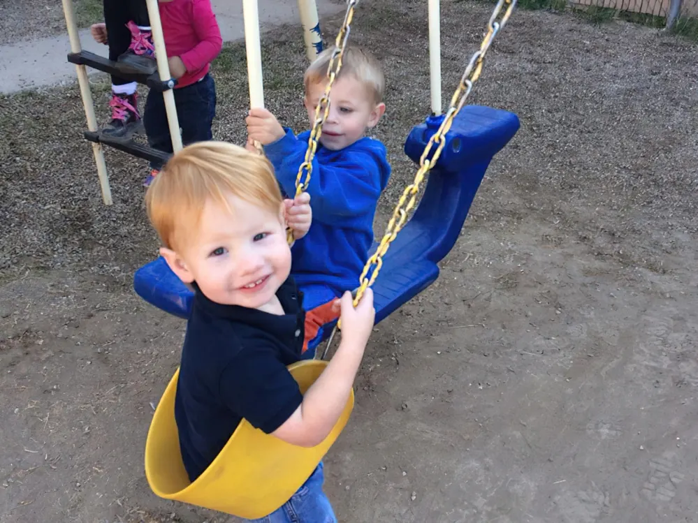Two toddlers riding a tandem swing glider with yellow and blue seats on a dirt playground