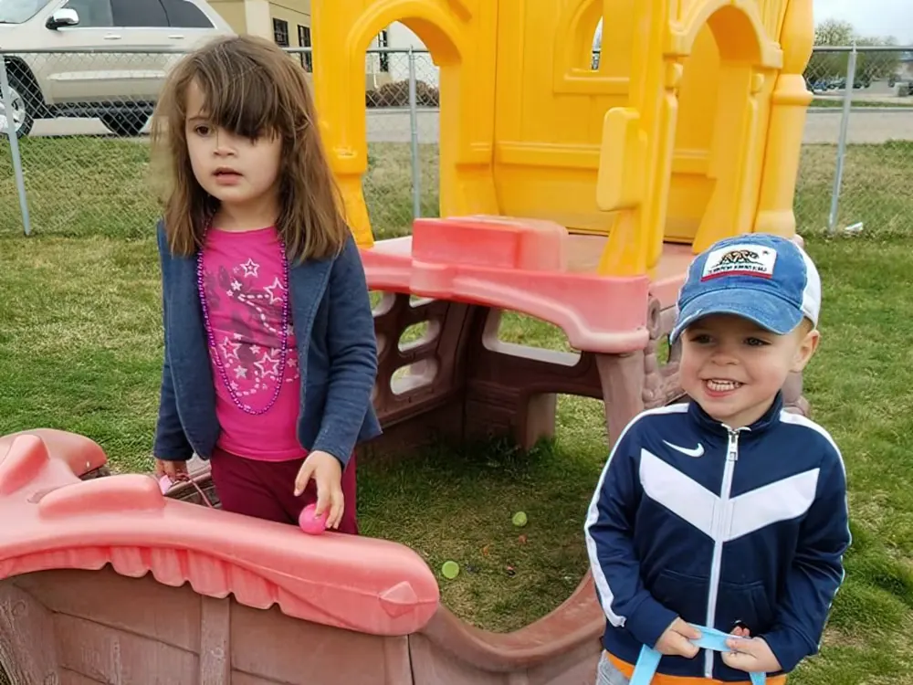 A girl in pink and a boy in a navy track jacket and cap beside a plastic playhouse outdoors