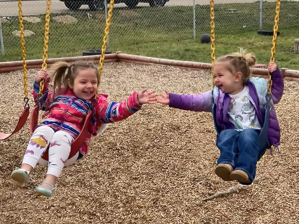 Two little girls on swings reaching out to hold hands over a wood-chip playground