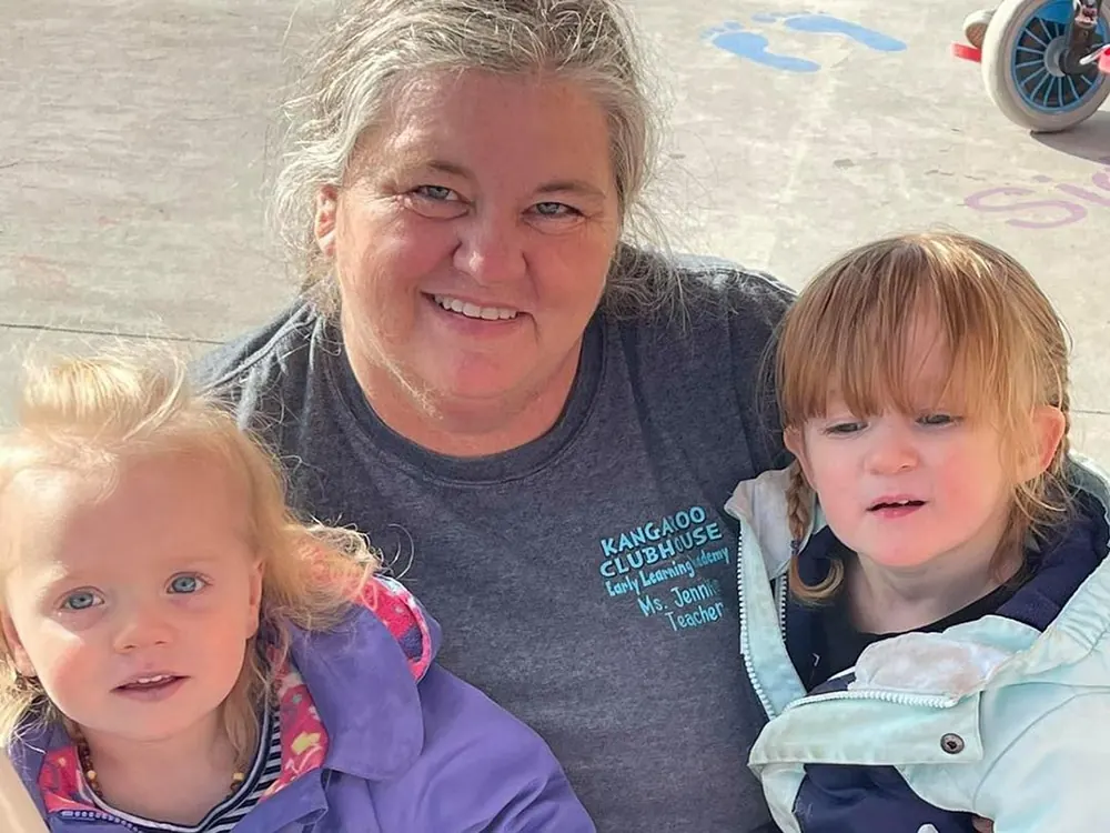 A smiling teacher in a Kangaroo Clubhouse T-shirt outdoors with two little girls in jackets