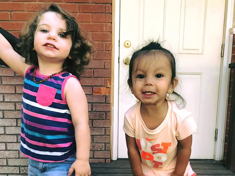 Two toddler girls standing on a step outside a brick building, one waving