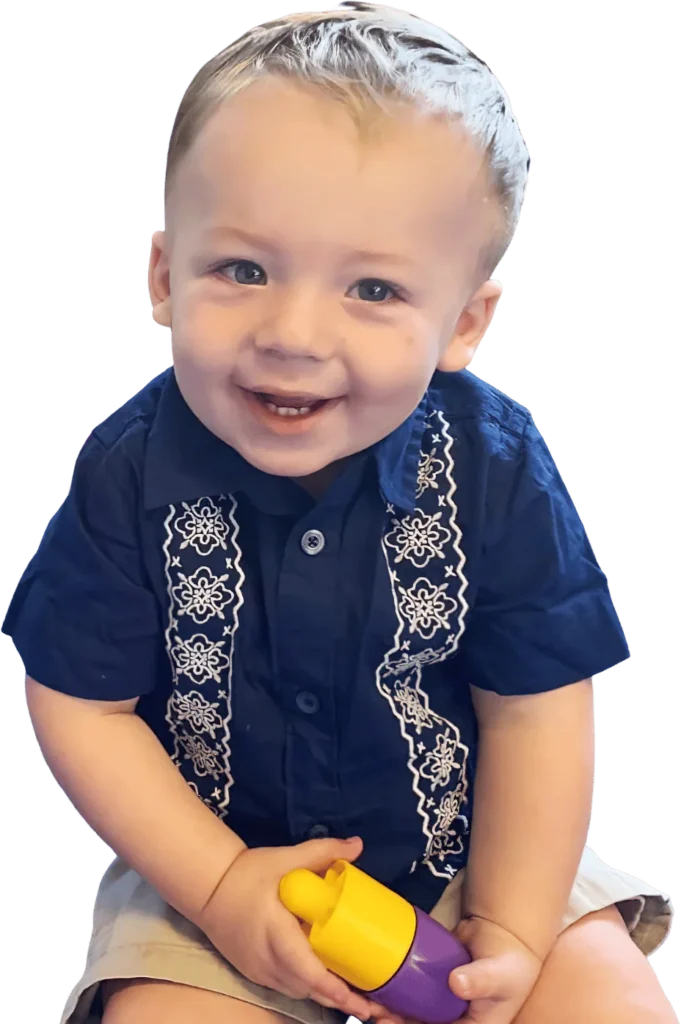 A grinning toddler boy in a navy embroidered shirt holding a yellow-and-purple toy