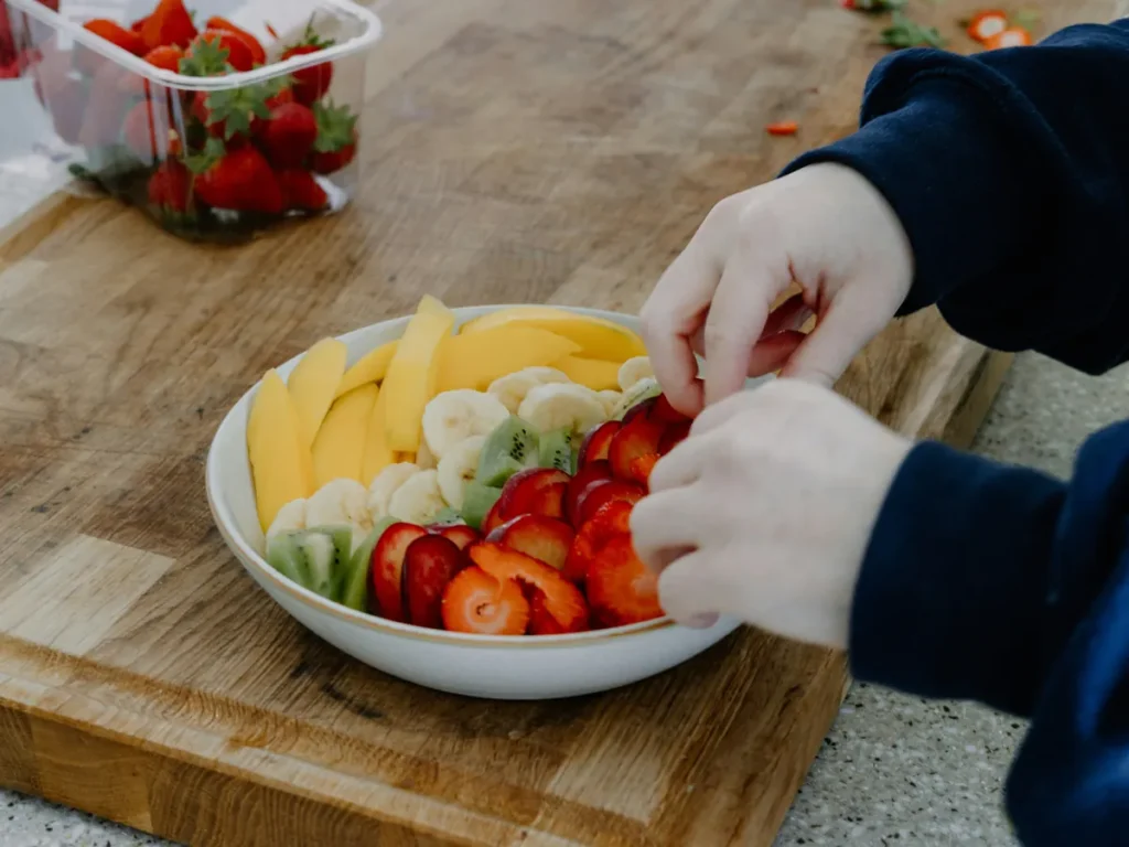 Hands arranging cut strawberries, kiwi, banana and mango in a bowl