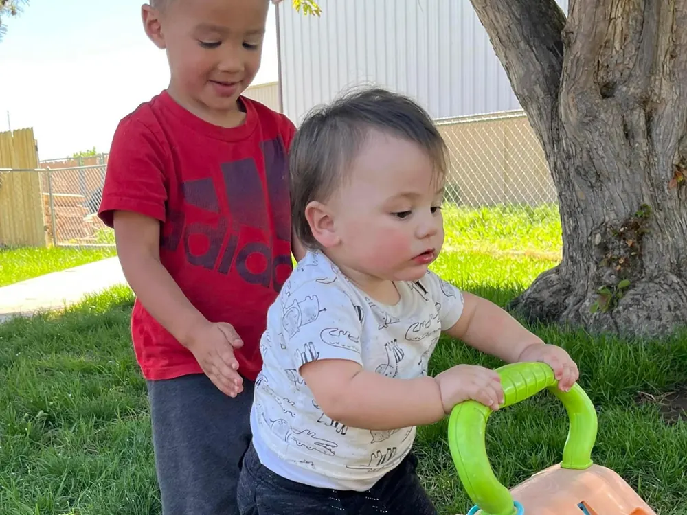A toddler pushing a ride-on toy with a green handle across the grass, an older boy beside him