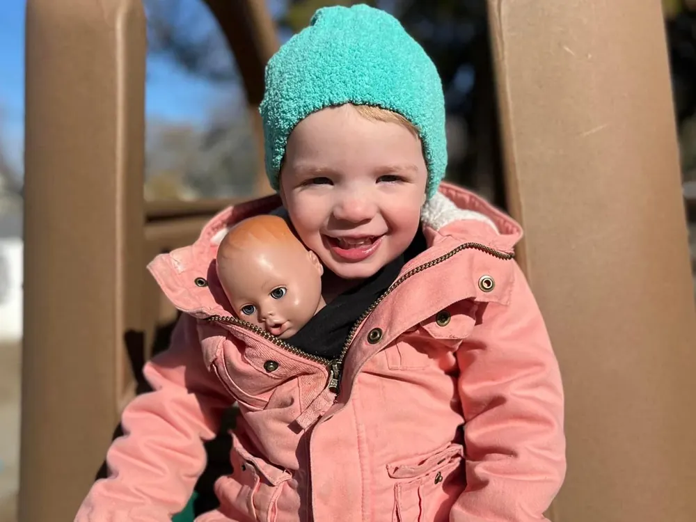 A smiling toddler in a teal knit hat and pink coat hugging a baby doll