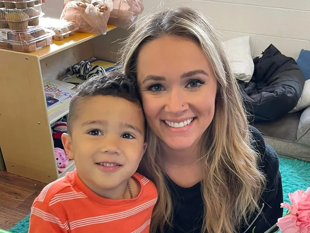 A smiling teacher with her arm around a boy in an orange striped shirt